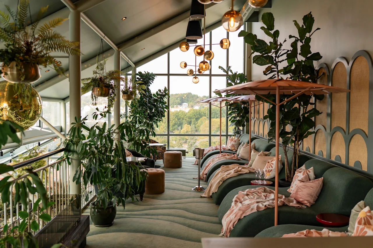 Rauhallinen, kasveilla täytetty oleskelutila, jossa on mukavat vihreät leposohvat, pehmeät peitot ja suuri ikkuna, josta avautuu luonnonkaunis näkymä.