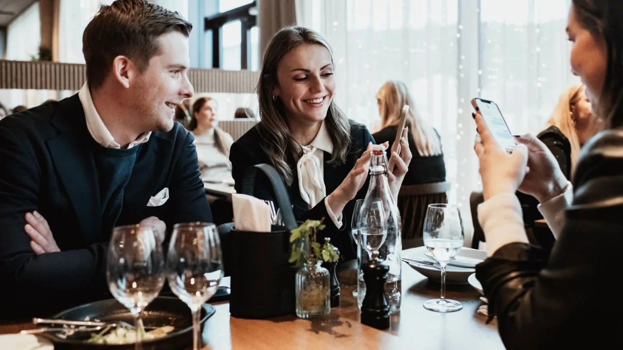 Tre personer umgås glatt vid ett restaurangbord, en visar sin telefon för de andra.