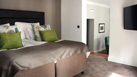 En ryddig seng med dekorative puter i et pent soverom, sollys strømmer inn fra et vindu, og en veggmontert TV på siden.