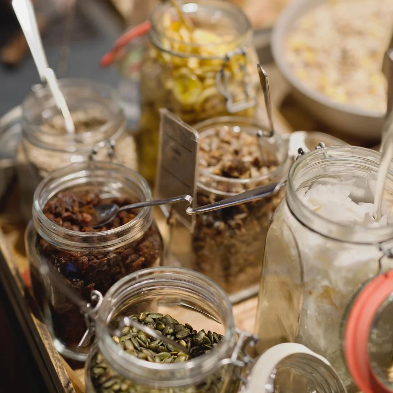Several glass jars with metal spoons, containing various dry foods, arranged on a wooden surface.