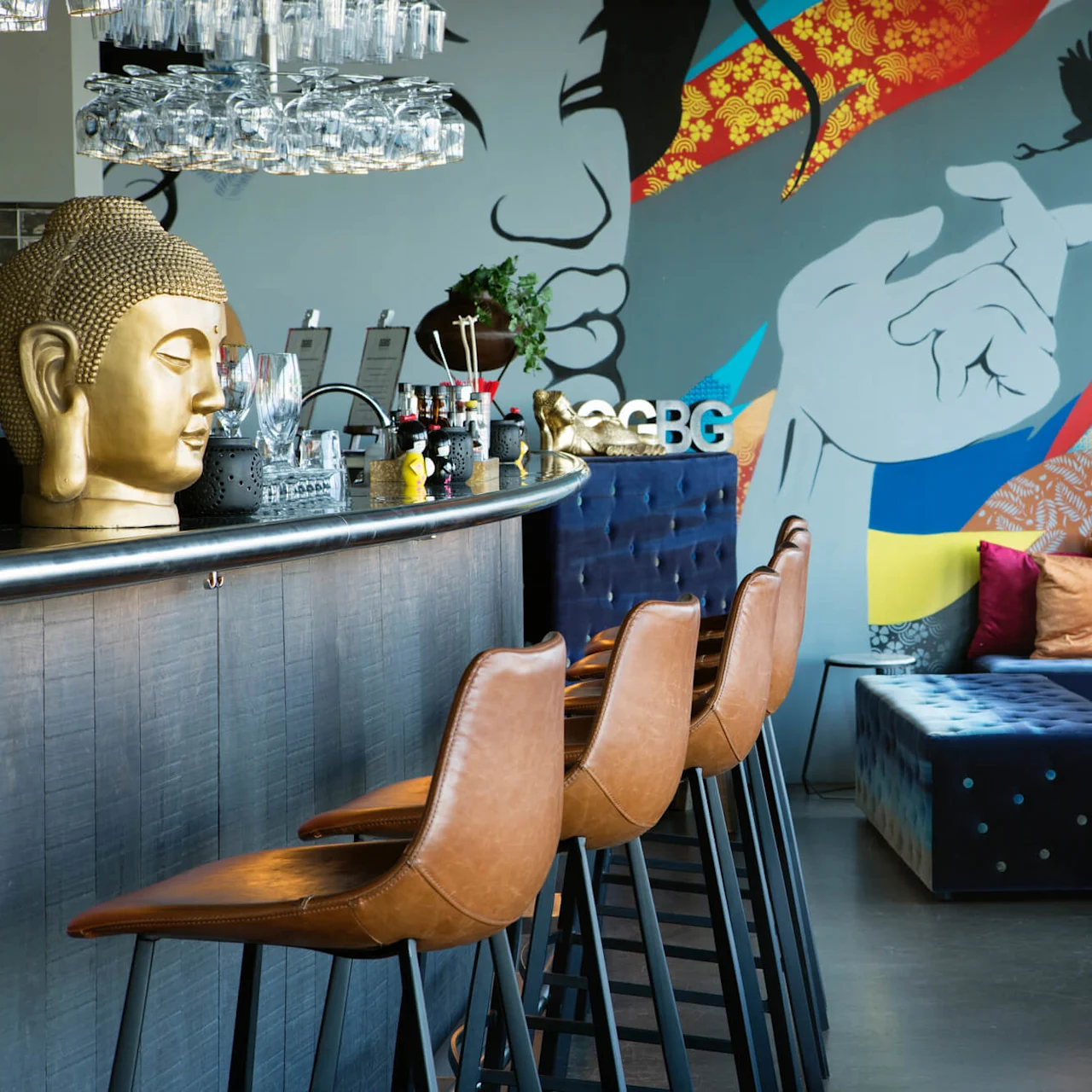 En bardisk med ett gyllene Buddhahuvud, läderpallar och hängande glas framför en färgglad mural med abstrakta mönster och fåglar. Texten "GBG" visas på en hylla.