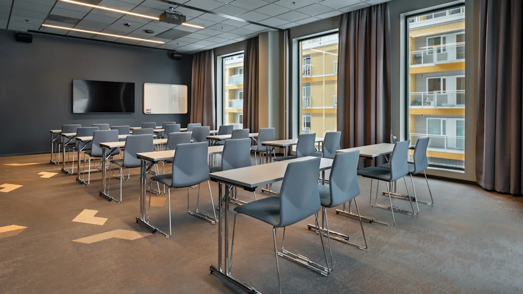 Rows of empty gray chairs face forward in a modern classroom. Large windows reveal an adjacent building. A wall-mounted screen and whiteboard are visible at the front.