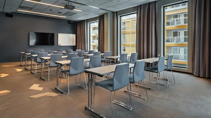Rows of empty gray chairs face forward in a modern classroom. Large windows reveal an adjacent building. A wall-mounted screen and whiteboard are visible at the front.