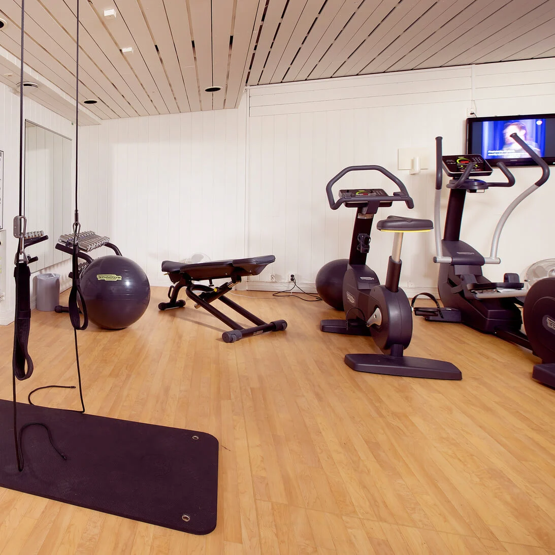 Træningsudstyr i et fitnesscenter med løbebånd, motionscykler og træningsbolde, tæt på plakater, der viser træningsrutiner.