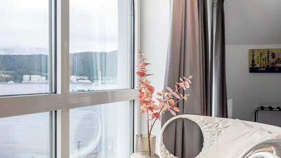 En modern stol med rund rygg står vid ett stort välvt fönster. En vas med rosa blommor står på ett litet bord bredvid, med utsikt över vatten.