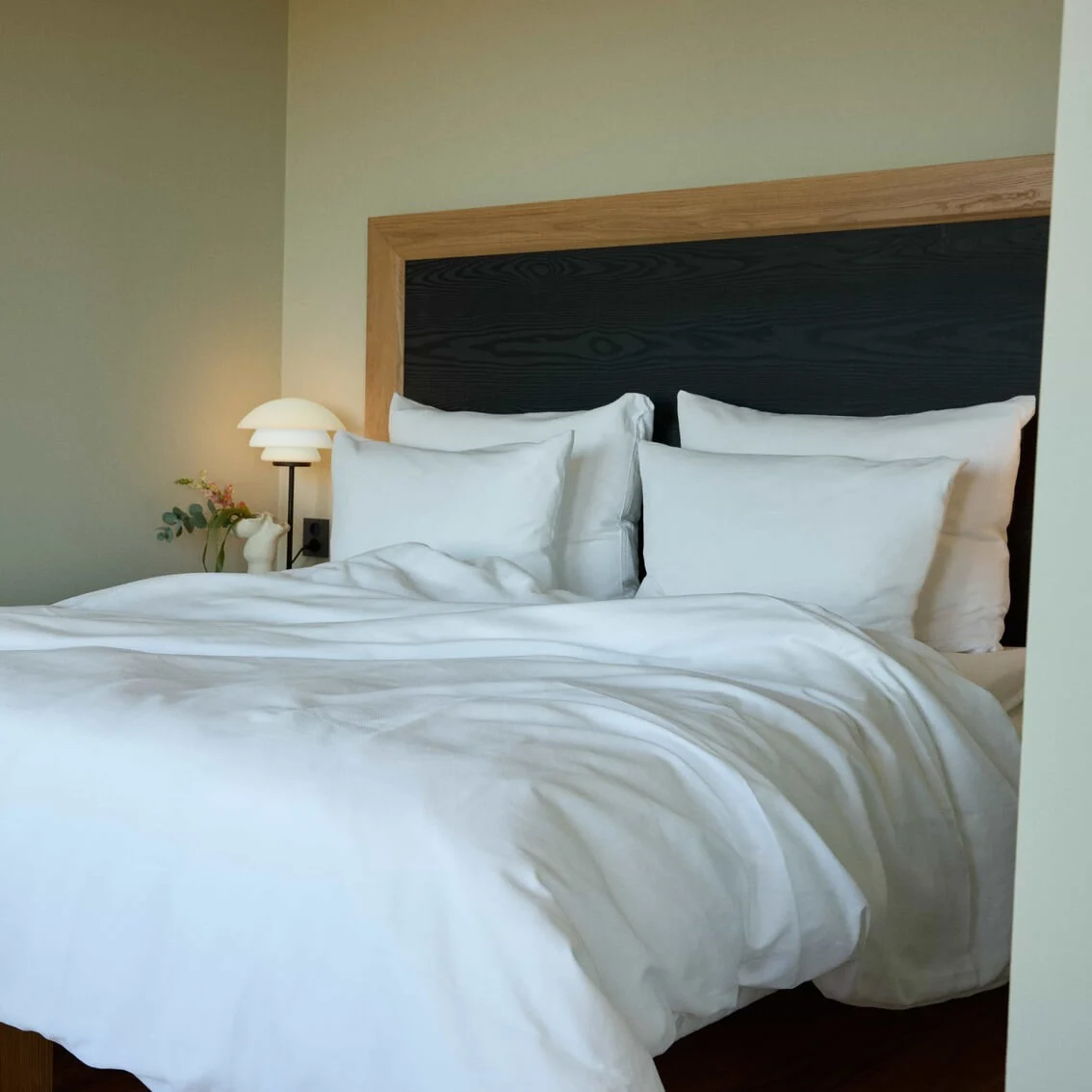 A neatly made bed with white linens sits against a wooden headboard. A bedside lamp glows softly on a nightstand with a small vase of flowers.
