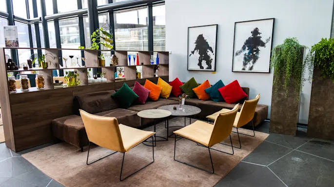 A seating area features a brown couch with colorful pillows and four tan chairs around circular tables. Shelves with plants and decor create a cozy atmosphere, alongside abstract artwork on white walls.