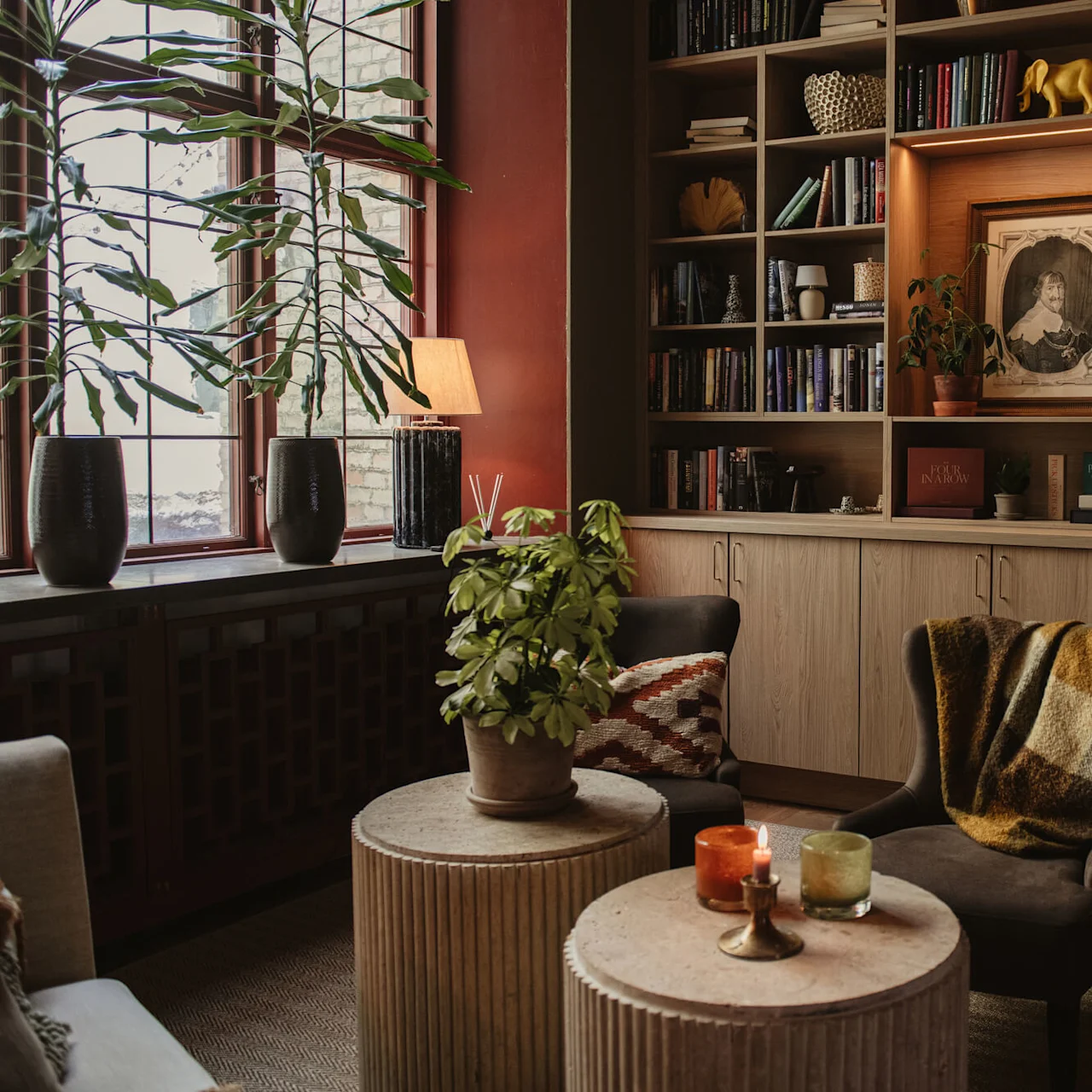 A cozy living room features two armchairs, round tables with candles, and large potted plants by a window. Bookshelves with books, decorative items, and a framed portrait fill the wall.