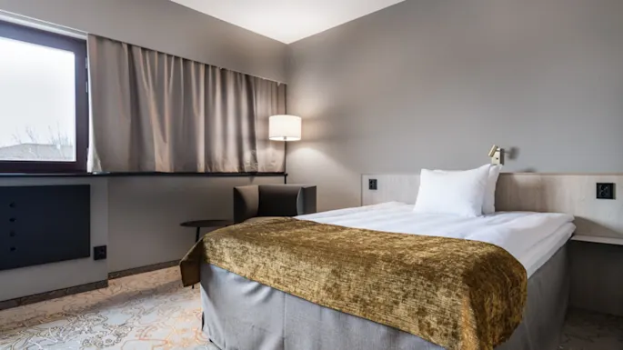 Seng står i et koselig, moderne hotellrom, dekorert med hvite laken og et gylldent sengeteppe. Ved siden av lyser en lampe opp de nøytrale veggene og gardinene.