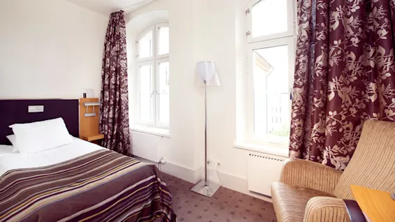 A neatly made bed with striped linens, adjacent to a window with floral curtains, in a well-lit, tidy hotel room.