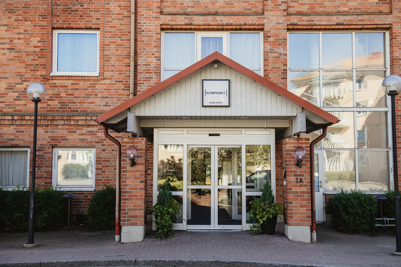 En välkomnande entré till en tegelbyggnad med glasdörrar, en skylt med HOME HOTEL och dekorativa växter. Adress 1A.