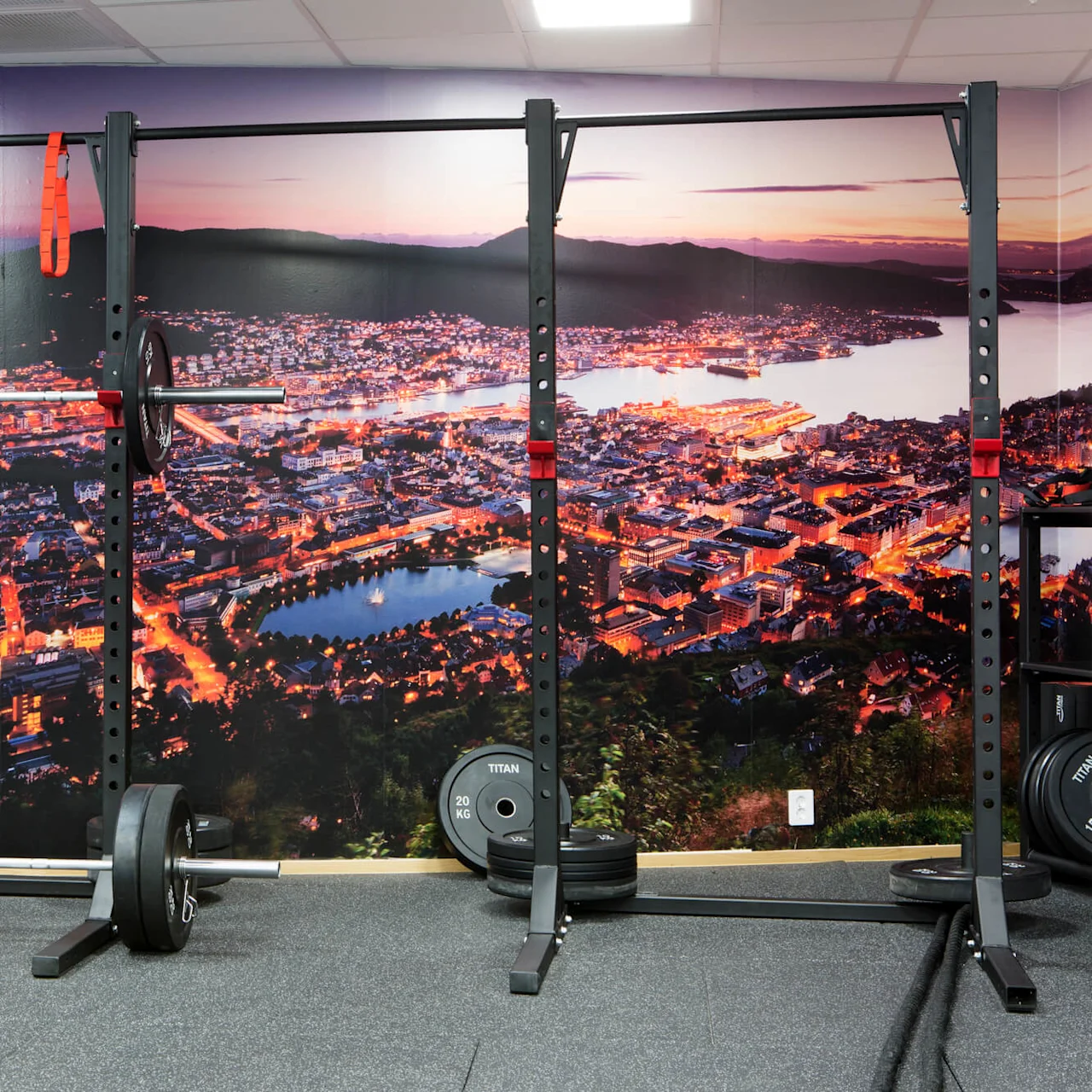 A weightlifting station featuring barbells with weights and a rack in a gym, set against a large cityscape mural. Plates labeled "TITAN 20 KG" are visible below.
