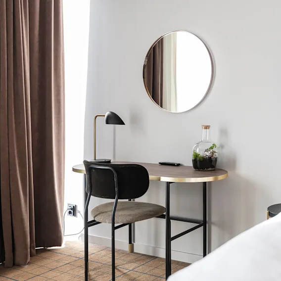 Desk supports a lamp, remote, and terrarium; placed beside a bed with a brown headboard. A round mirror hangs above in a room with brown curtains and carpet.