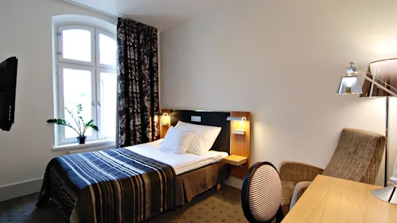 Cozy hotel room; a neatly made single bed with striped bedding rests against a wall, accompanied by a desk, chair, and window with a curtain; serene and tidy environment.