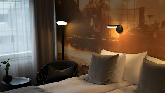 A neatly made bed with gray pillows is illuminated by a wall lamp against a backdrop of a warmly lit mural. An armchair and side table are nearby.