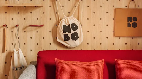 A modern living space with a red sofa and a wooden pegboard wall displaying bags, shelves, and a lamp. 