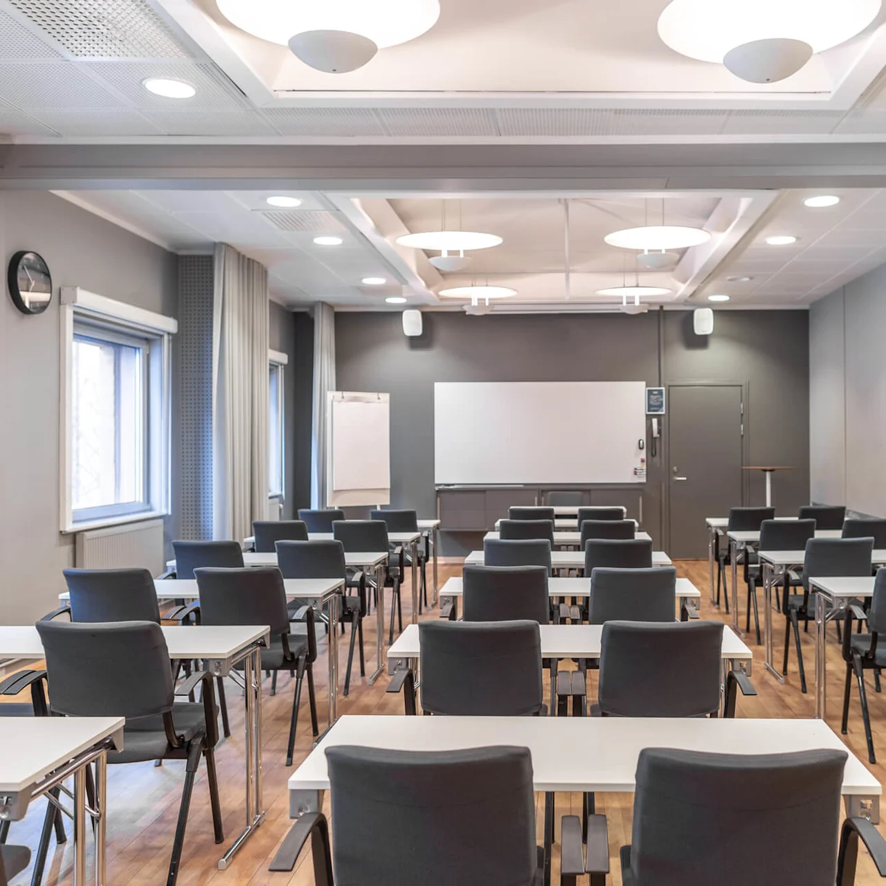 Rows of empty desks with chairs face a whiteboard in a well-lit, modern classroom. Windows and a flip chart are on the left side.