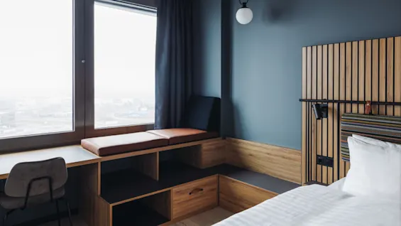 Window seat with cushions in a modern room, next to a large window. Wood paneling and dark curtains enhance the minimalist decor, with a chair and bed nearby.
