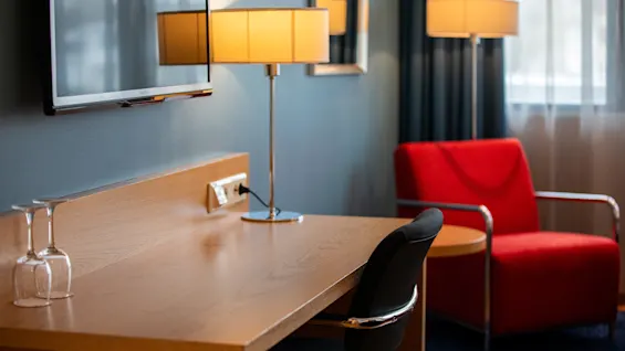 Et skrivebord i tre med to opp-ned glass ved siden av en svart stol, i et rom med veggmontert TV, lampe, rød lenestol og gardiner. Tekst: "Philips".