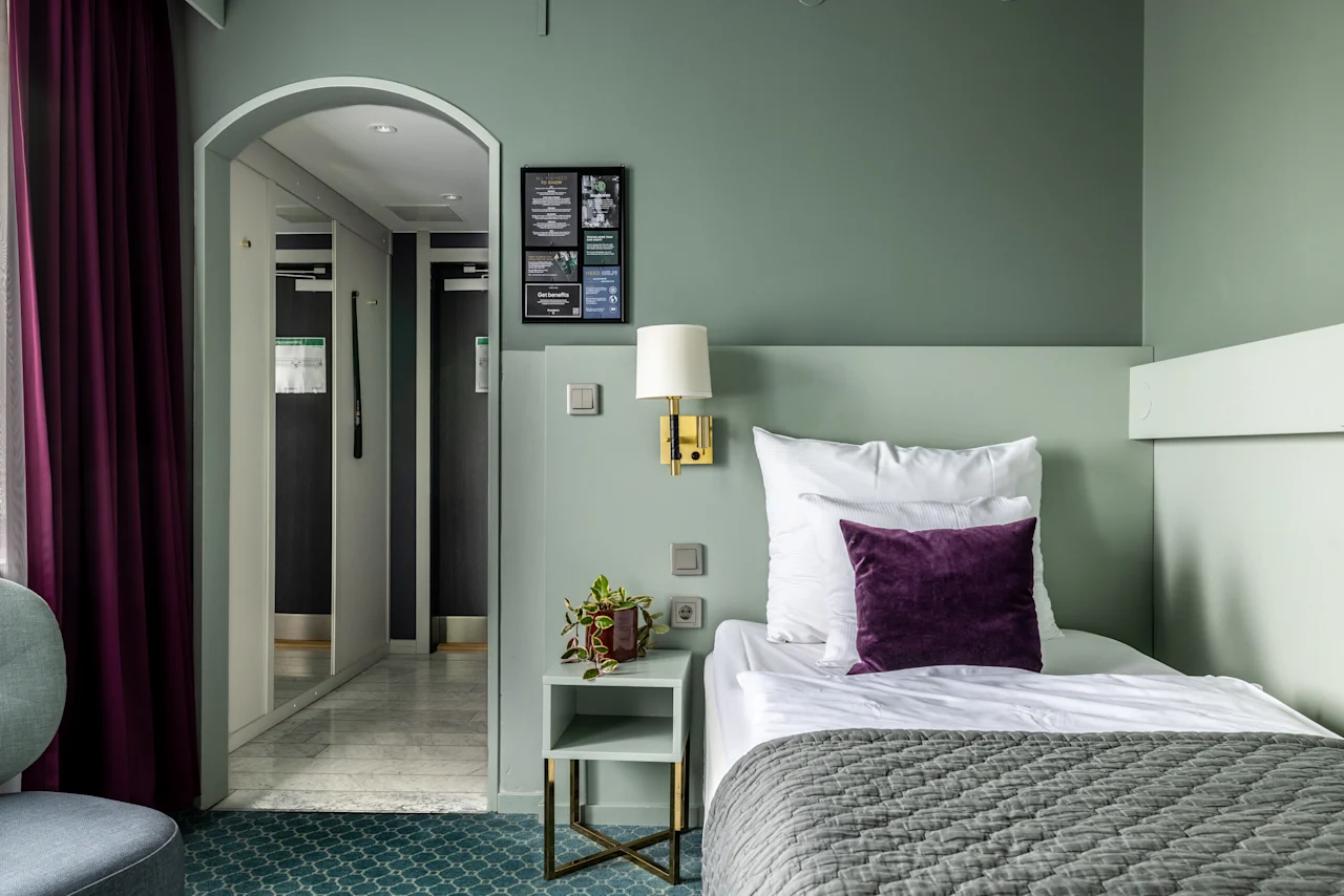 Kodikas hotellihuone, jossa on yksi vuode koristeltuna valkoisilla ja violetilla tyynyillä. Pehmeät vihreät sävyt luovat rauhoittavan ilmapiirin, jota täydentää pieni kasvi vieressä. Taustalla käytävä tuo syvyyttä.