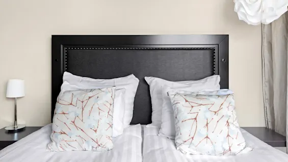 A neatly made bed with patterned pillows, against a plain headboard, beside a white lamp and a decorative light fixture.