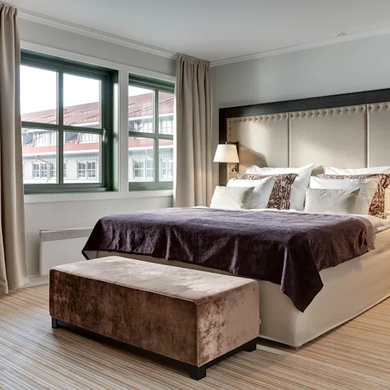 A bed with multiple pillows and a dark blanket stands against a cushioned headboard. Two lamps adorn the sides. Windows with beige curtains overlook neighboring rooftops.