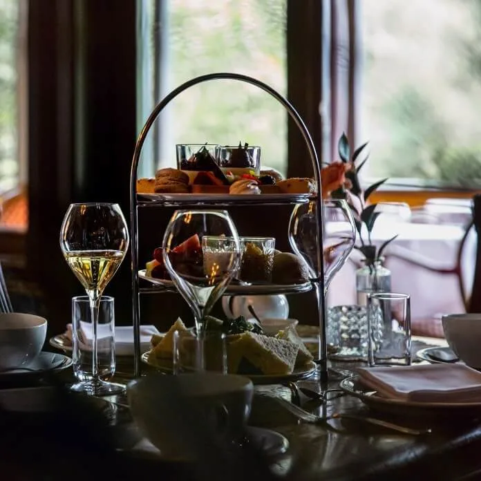 Afternoon tea set with tiered trays holding desserts and sandwiches, alongside wine glasses, cups, and plates on a dark wood table, in an elegant, softly lit dining room.