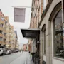 En travl europæisk gadescene med et HOME HOTEL-banner, historisk arkitektur og folk, der nyder en gåtur eller cykeltur.