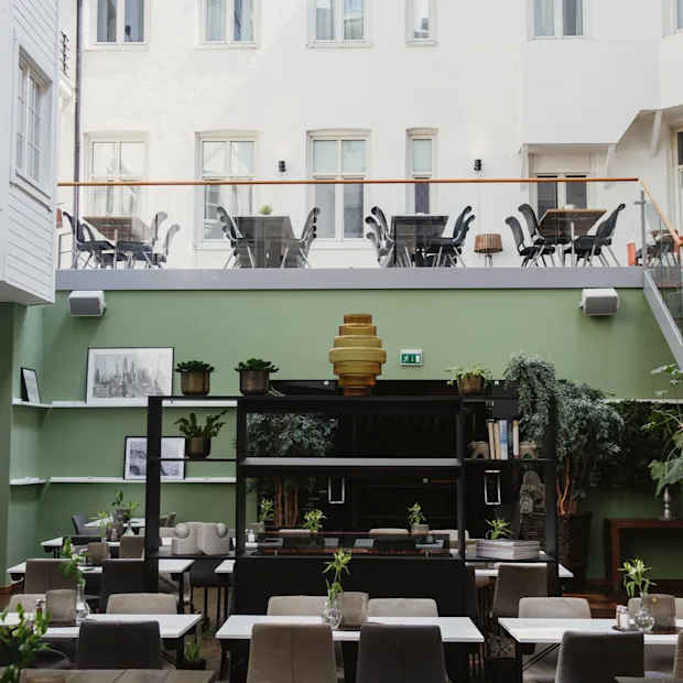 Restaurangbord med växter; omgivande grön inredning och konstverk skapar en modern, luftig atmosfär. Utomhusplatser på övervåningen syns genom en glasbarriär.