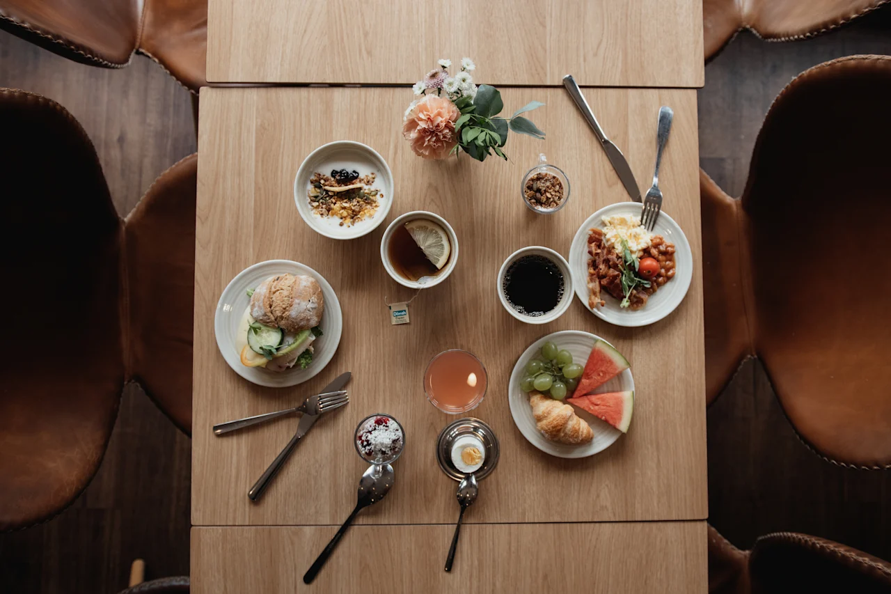 An inviting breakfast spread on a wooden table, featuring a variety of dishes like a sandwich, yogurt, fruit, and hot drinks.