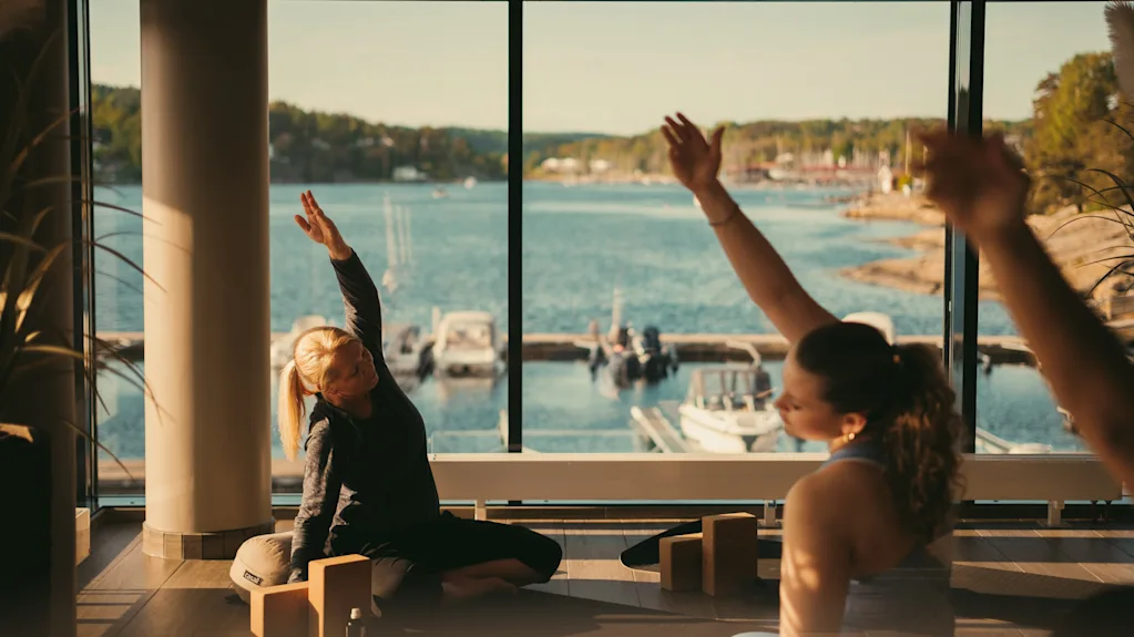 Folk der strækker sig indendørs med en naturskøn udsigt over en sø og både uden for et stort vindue, nyder et fredeligt øjeblik.