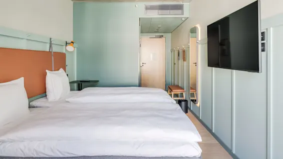 En pænt redt seng med hvidt linned står i et minimalistisk hotelværelse. Et vægmonteret fjernsyn og en trædør er synlige i baggrunden. Bløde grønne vægge tilfører ro.