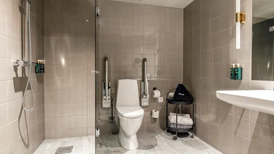 Toilet med sikkerhedsrækværk i et moderne badeværelse, med en bruser til venstre, beige flisevægge, en hvid håndvask og hylder med toiletartikler til højre.