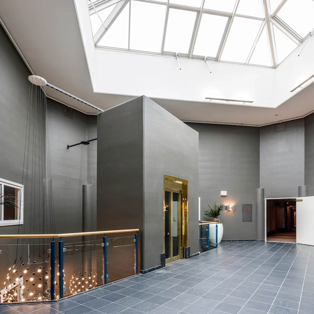 A glass-ceilinged atrium with gray walls and tiled floor features a central golden elevator. A modern chandelier hangs nearby, while potted plants decorate the space.