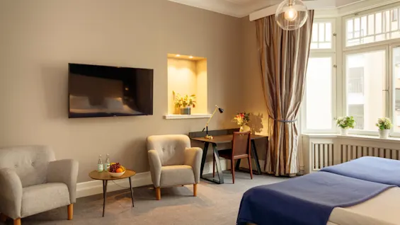 Ett mysigt rum har två grå fåtöljer och en vägghängd TV. Ett litet bord med frukt och flaskor står mellan dem. Ett skrivbord med lampa och blommor är i närheten, bredvid stora fönster med gardiner.