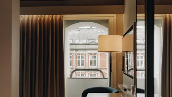Ett mysigt rum med skrivbord och lampa, med utsikt över stadens byggnader genom ett välvt fönster.