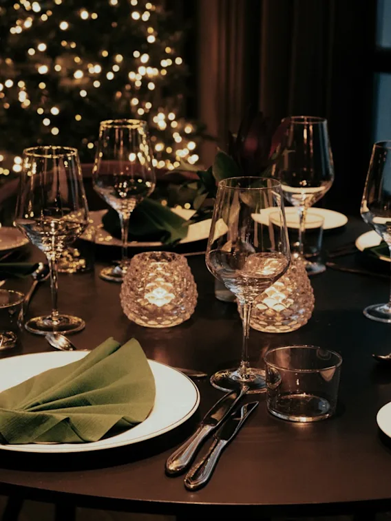 Et elegant dekket rundt middagsbord med vinglass, grønne servietter, levende lys og uskarpe juletrelys i bakgrunnen.