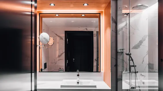 A modern bathroom features a rectangular mirror with backlighting, a white sink beneath it, a wall-mounted magnifying mirror to the side, and a glass-enclosed shower area with marbled tiles.