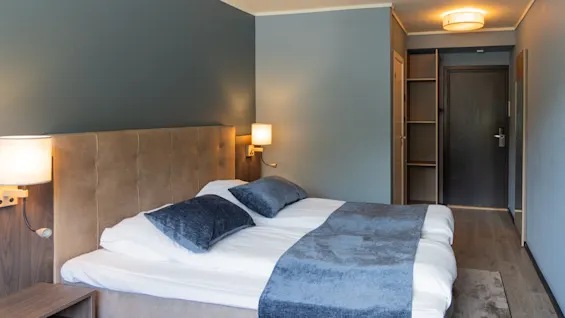 En prydligt bäddad dubbelsäng med blåa detaljer står i ett litet, modernt hotellrum med blågrå väggar, trägolv, ett sängbord med lampor och en dörröppning som leder till en korridor.