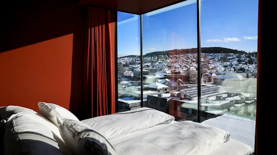 En seng med hvide puder er placeret foran store vinduer med udsigt over et snedækket bylandskab under en klar blå himmel.