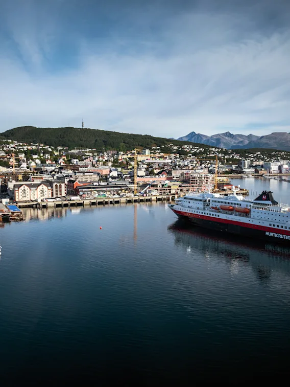 Et stort passasjerskip seiler gjennom en havn med en by som ligger inntil åser og fjell under en blå himmel.