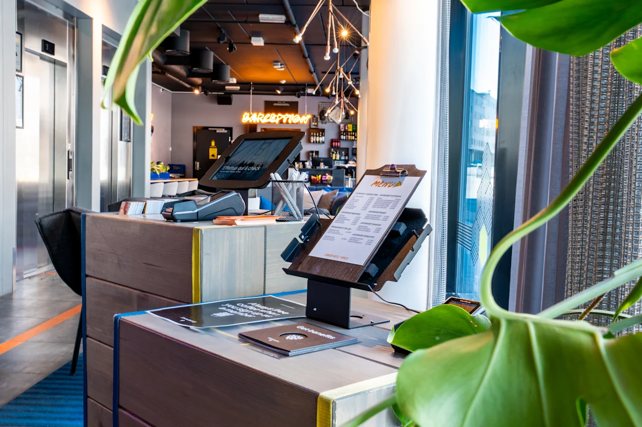 En digital indtjekningskiosk med skærm og clipboard står på et træbord i en moderne hotellobby. I nærheden lyser et neonskilt med teksten "RECEPTION" over receptionen.