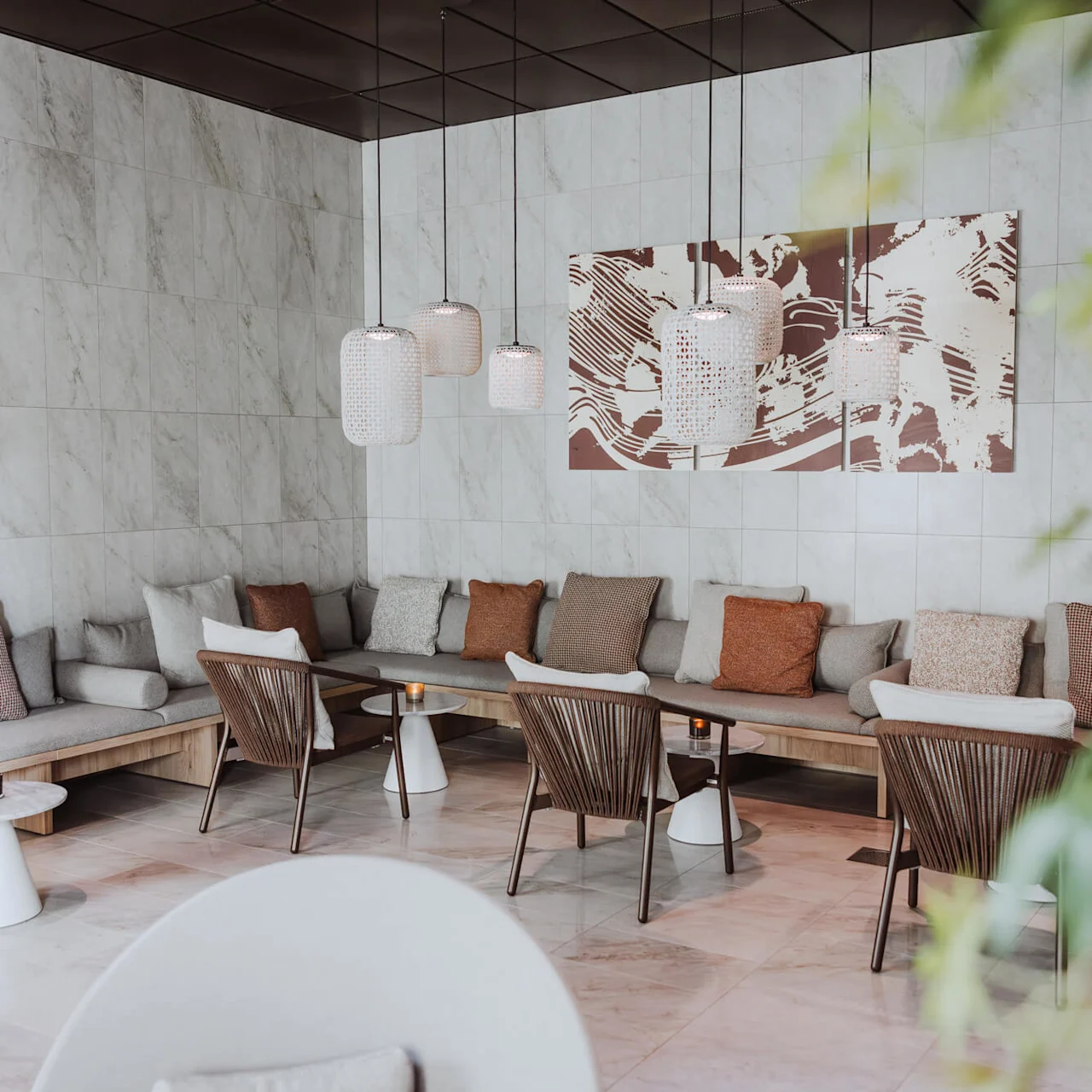 Sittområde med kuddar på bänkar och stolar runt små bord under hängande lampor, i en modern, minimalistisk lounge med kaklade väggar och abstrakt konst.