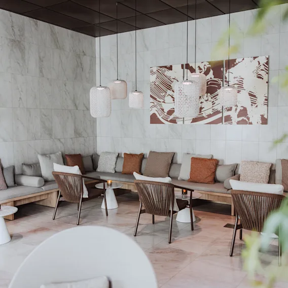 Sitteområde med puter på benker og stoler rundt små bord under hengende lamper, i en moderne, minimalistisk lounge med flislagte vegger og abstrakt kunst.