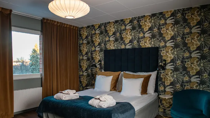 En seng med pent plasserte håndklær står i et rom med blomstrete tapet, fløyelsgardiner og en komfortabel stol, opplyst av en taklampe.