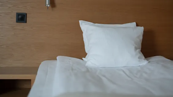 A neatly made bed with white pillows rests against a wooden wall. A small wall light and a power outlet are visible. The setting is minimalist and modern.