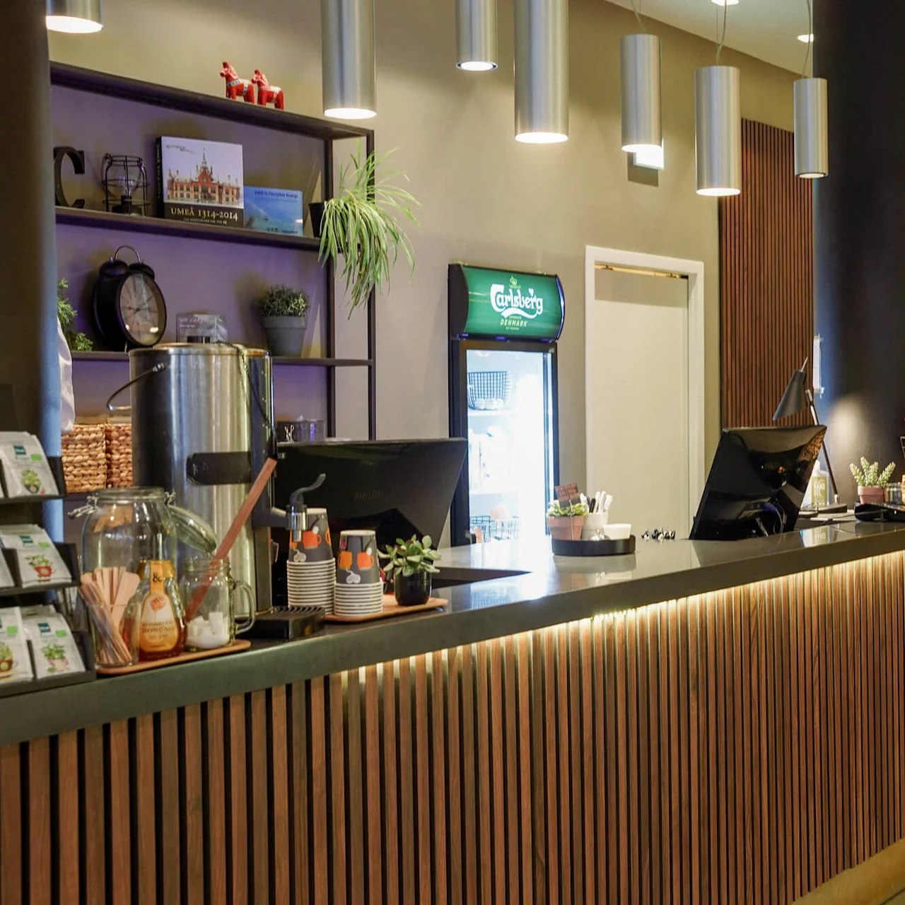 En moderne hotelreceptionsdisk med snacks, planter og en kaffedispenser, sat op imod en baggrund af afdæmpet belysning og nutidig indretning.