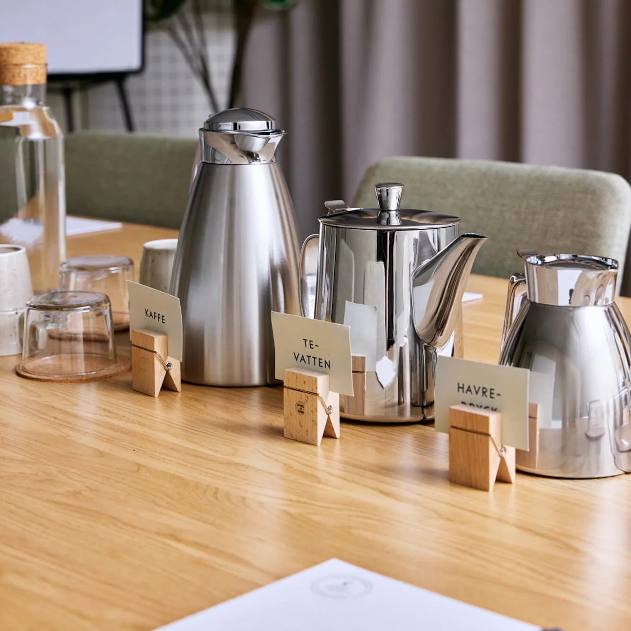 Silver thermoses and cards labeled "KAFFE," "TEVATTEN," "HAVREVÄXT" on a wooden table with glasses and mugs, surrounded by chairs and a plant in a meeting room.