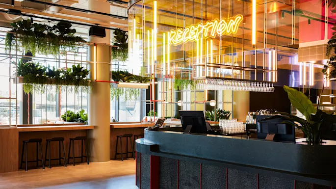 Resepsjonsskranke med planter og neonskilt som sier "RECEPTION"; livlig moderne miljø med hengende glass og krakker nær vinduer med naturlig lys.