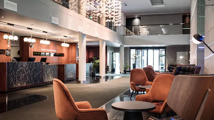 A reception desk with marble panels is attended and illuminated by modern lights; surrounded by plush chairs and tables in a spacious, stylish hotel lobby. Sign reads: "Quality Hotel Reception."